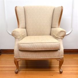 Wingback Armchair: A wingback armchair. This chair features a cherry-finished wooden frame with foliate carvings. It is upholstered in a swirl-patterned cream color fabric. It sits on cabriole style legs with carved pad feet.