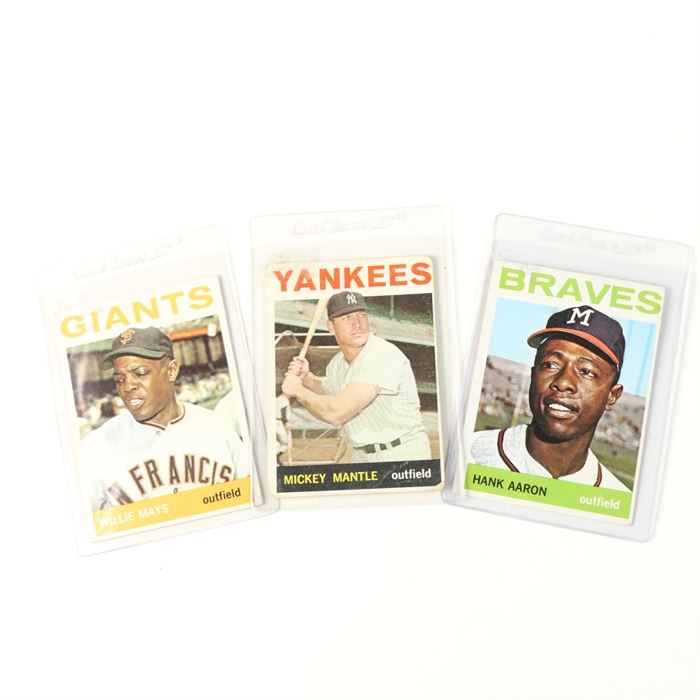 1964 Willie Mays, Mickey Mantle, and Hank Aaron Topps Cards: A collection of 1964 Topps baseball cards. Featured are no. 150 of Willie Mays for the San Francisco Giants, no. 50 of Mickey Mantle for the New York Yankees, and no. 300 of Hank Aaron for the Milwaukee Braves. Each card shows a color depiction to the front, with athlete statistics to the verso, and is held in a protective sleeve.