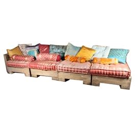 Teak Wood Outdoor Sectional Couch: A teak wood outdoor sectional couch. This set includes four square back pieces with square seat resting on bracket feet. One piece features a tall armrest. This set includes decorative seat cushions and accent pillows. There are no visible maker’s marks or labels on these pieces.