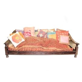 Tibetan-Style Couch: A Tibetan-style couch. This wooden couch features a slatted back and seat with sloping arms and turned arm supports. It rests upon bun feet and includes a red rectangular seat cushion with gilt trim and ornamentation. A group of throw pillows in shades of red, pink, blue, yellow, green, and orange are included. See matching items 17LAX108-002, 17LAX108-004, and 17LAX108-007.