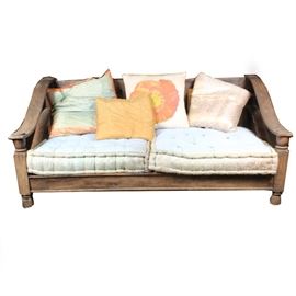 Tibetan-Style Love Seat: A Tibetan-style love seat. This wooden seat features a slatted back and seat with sloping arms and turned arm supports. It rests upon bun feet and includes two rectangular blue seat cushions with a floral border. Four throw pillows in shades of blue, gold, yellow, red, and white are included. See matching items 17LAX108-002, 17LAX108-003, and 17LAX108-004.