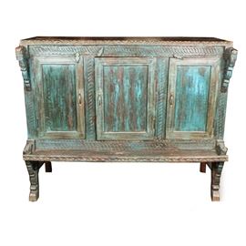 Tibetan-Style Cabinet: A Tibetan-style cabinet. This wooden cabinet has a distressed blue-painted finish and features three cabinet doors with brass tone pulls and wooden swivel latches. The piece contains two interior shelves and a protruding exterior shelf below the doors. The piece is embellished with carved stylized dragons in relief to the four front corners and incised geometric designs throughout. It rests upon carved legs with metal lateral stretchers.