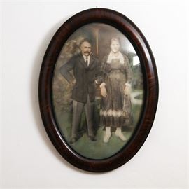 Antique Hand Tinted Photograph: A framed hand tinted antique photograph of a man and woman. The man’s expressive pose and the woman’s steady gaze are augmented by the addition of naturalistic coloring. The photograph is framed in an oval wooden frame under domed glass.