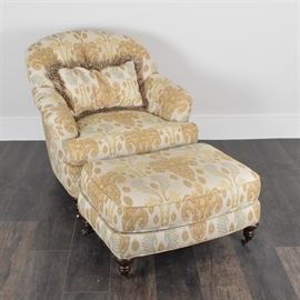 Barrel Style Chair with Ottoman by Lee: A barrel style chair and ottoman by Lee. The chair and ottoman are wood with a walnut finish and feature a chintz cloth upholstery in a cream color with a brown and green floral design and cloth piping accents. The chair has a rolled crest rail and a curved and tufted back and arms with a loose cushioned seat and tapered legs on caster feet. The matching ottoman has a rectangular shape and features tapered legs on casters. The chair and ottoman are labeled “Lee Industries”.