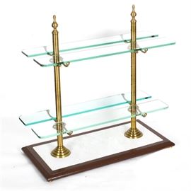 Brass and Glass Patisserie Serving Stand: A brass and glass patisserie serving stand. This stand features a two tier design with glass shelves and brass connectors displayed to a marble and wooden base with beveled edges. This stand displays no visible brand labels.