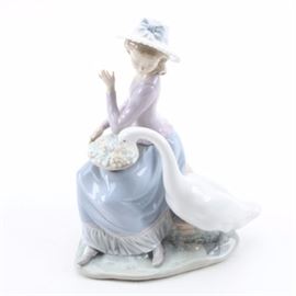 Retired Lladro Figurine "Goose Trying to Eat" #05034: A retired Lladro porcelain figurine, titled Goose Trying to Eat. This figurine is #05034 and depicts a woman wearing a flowing dress and hat with a bow, who sits on a rock while twisting away as a lone goose tries to pick at a plate of grapes resting in her lap. The surface has been painted in light pastel tones. To the underside is stamped manufacturer information.