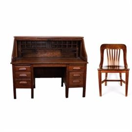 Vintage Roll Top Desk With Matching Chair: An antique roll top desk with matching chair. This pair includes an antique desk and chair made of solid oak. The desk features five drawers with three on one side and two on the other and rows of slots and cubby holes for office accessories. There is a pull-out writing surface on either side above the column of drawers. The front of the desk rolls down. This antique piece is made with intricate dovetail joints and has a varnish finish. There is a single brass accent piece where the roll top latches and locks. There is no key included. This desk is circa 1900. Also included is a matching chair. It has a curved seat back with a slotted design. It too is made of solid oak and has a smooth and polished finish. Both these pieces are unmarked.
