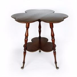 Victorian Parlor Table: A Victorian parlor table. This Victorian parlor table has a clover design. It features a large table surface and smaller matching shelf beneath. The legs have a spindle design with claw and ball feet. There is metal accent trim around both the table surface as well as the lower shelf.