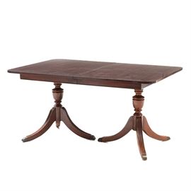 Duncan Phyfe Style Mahogany Dining Table: A mahogany Duncan Phyfe style dining table. This table has a rectangular top with round edges, straight apron, and a center leaf extension. It stands on two turned pedestal bases with splayed, reeded legs and brass end caps.