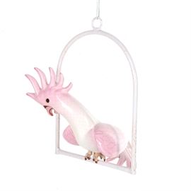 Hanging Pink Ceramic Cockatiel Decor: A hanging pink ceramic cockatiel figurine. This figurine depicts a cockatiel perched on a swing, with its pink crest raised up, looking down at something below. It has black eyes, painted feathers, and red nails on its feet. Marked to the underside is “Coral Mex”.