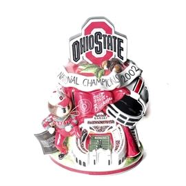Christopher Radko Ohio State 2002 Cookie Jar: A Christopher Radko ceramic cookie jar commemorating the Ohio State University 2002 National Championship football season. The base of the figural jar has numerous OSU Buckeye related items including Brutus, the Horseshoe, football helmet, drum major’s hat, bass drum, megaphone, and the lyrics to verse 1 of “Carmen Ohio”. The top has the OSU “Block O” emblem and a ribbon with the years of past championship seasons. It is marked Christopher Radko and “The Perfect Season” on the bottom. The jar comes with its original box.