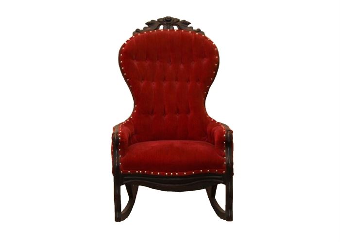 Antique Victorian Rocker: An antique walnut Victorian rocker that features carved and molded detailing, and the upholstered backrest and seat are covered with a red velvet style tufted upholstery bordered by nail heads. The chair has carved front legs and curved back legs attached to rockers.