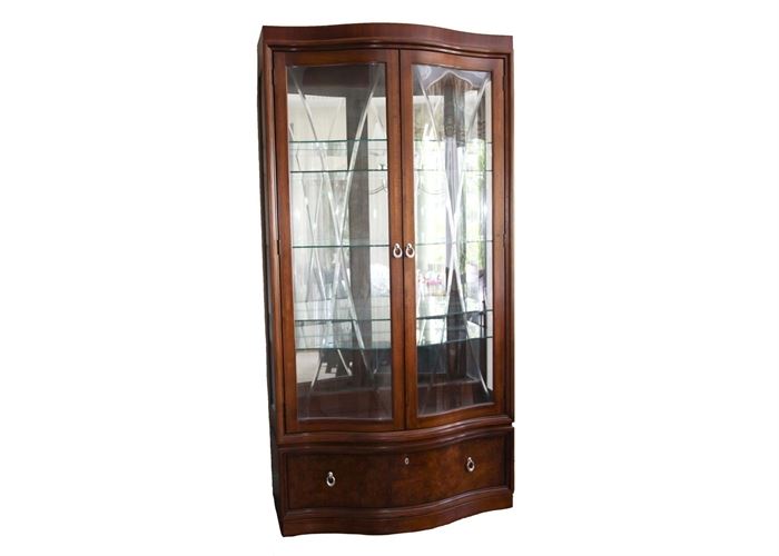 Thomasville Curio Cabinet: A Thomasville lighted curio cabinet. This mahogany finished cabinet has a serpentine front with beveled V-grooved bowed glass cabinet doors. The interior space features glass shelves, glass sides and a mirrored back. A burled finished bow front storage drawer beneath the cabinet doors has dovetail joints and silver-tone loop pulls.