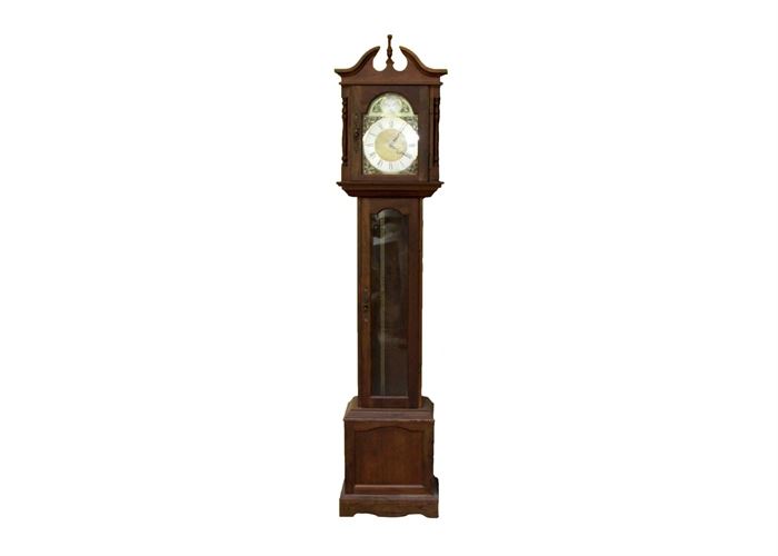Tempus Fugit Grandfather Clock: A Tempus Fugit grandfather clock. This clock has a walnut tone cabinet with base molding, a decorative base panel and elongated trunk where a door with a glass panel is located to view the brass pendulum and chains and weights. It has a broken pediment with a center finial. The clock face has a brass tone finish, black Roman numerals on a silver tone ring. Filigree scrolled detail is applied surrounding the clock face. “Tempus Fugit” is labeled above the face and "Emperor"is marked to the clock face.