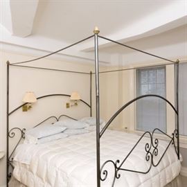 Metal Canopy Bed: A metal canopy bed. This piece features a dark gray finish, with brass-toned accents. To the top are tubular stretchers connecting the four posts, with brass-toned pineapple finials to the tops. The headboard and footboard have arched tubular rails connecting their respective posts, with scrolling metal work below. The side rails feature scrolling metal accents, as well.