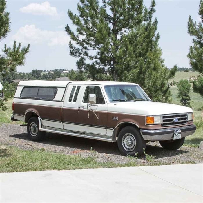 1990 Ford F250 XLT Lariat Pickup Truck: A 1990 Ford F250 XLT Lariat 2-door pickup truck with a Leer “Challenger” topper. At the time of cataloging the odometer reading was 119,172 miles. The VIN is 1FTHX25GXLKA40076. The 2-door truck is equipped with a V8 gasoline powered 7.5-liter engine, automatic transmission and rear wheel drive. The body is white with wide brown stripes on the sides and has Owens Classic XLR200 running boards along the sides. The truck is also equipped with air conditioning, and an AM/FM radio with a tape deck. The interior has tan carpet with tan cloth seats in front, and tan vinyl upholstery on the rear bench seat. Includes a trailer hitch and ball.