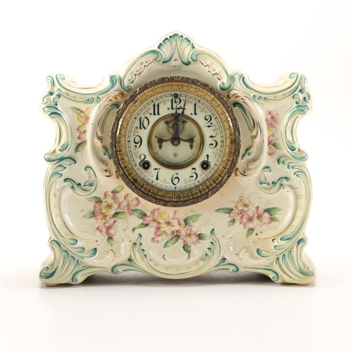 Ansonia Porcelain Mantel Clock: An Ansonia porcelain mantel clock. This ornate ceramic mantel clock features a scalloped top with scrolled sides, flowing floral accents and a molded flower accented base in a green hue. The clock face is white with black Arabic numbers and is framed in a textured brass frame with a removable brass back-plate to access mechanical works. The original key is included.