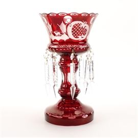 Bohemian Cranberry Glass Lustre: A Bohemian cranberry glass lustre. Also known as a girandole, this piece features a flared rim with a scalloped rim decorated with an etched pattern set upon a column shaped stem on a round base. The whole cased cranberry etched and cut to clear with geometric and landscape scenes. The sconce has cut colorless crystal notched spear form drops attached. The candle holder is unmarked.