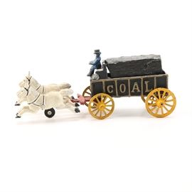 Cast Iron Horse and Coal Buggy: A cast iron horse and coal buggy. This cast iron toy features two white horses which are pulling a black buggy with gold tone lettering that reads “COAL” and rests on yellow wheels. The buggy features a man wearing blue and a black top hat and a black rectangular object depicting coal. The buggy is marked “Taiwan” to its underside.