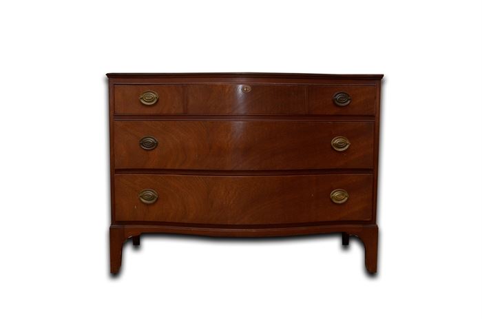 Hepplewhite Style Mahogany Chest of Drawers: A Hepplewhite style mahogany chest of drawers. This chest features a bow front with three drawers. Each drawer features bail pulls with oval backplates, with the top also having a center escutcheon. It sits on bracket feet. A partial “Genuine Mahogany” label is present to the interior of a drawer.