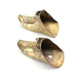 Brass Slipper Wall Sconces: A pair of brass wall sconces. In the form of slippers, these features curved pointed toes with raised figural accents with scrolled headdresses. There are triangular holes on the back for mounting. No discernible maker’s mark was found.