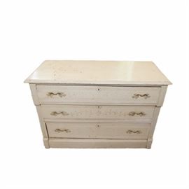 Eastlake Style Painted Chest of Drawers: An Eastlake style dresser with a cream painted finish. This piece has a rectangular top above three stacked drawers with incised floral designs and gilded foliate pulls. The top drawer protrudes slightly above the others with corresponding protruding stiles to either side. The piece stands on a molded base.