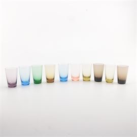 Ten Colored Shot Glasses: An assortment of ten colored shot glasses. These glasses have thick bases and slightly tapered straight sides. They come in colors including purple, blue, green, amber, light peach, and brown.