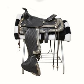 Simco Parade Saddle: A Simco parade saddle. Featured is a black show saddle, decorated with silver tone diamonds throughout the front and back jockey. The saddle shows a low cantle, decorative saddle straps, and covered stirrups. Also included is a black and white Corona saddle pad, and a metal saddle stand.