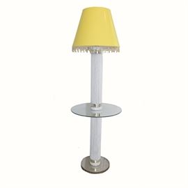 Floor Lamp With Small Glass Table: A floor lamp with a small glass table. This lamp has a yellow deep empire style shade with glass beads hanging from the edge, and a thick reeded body which supports a small glass table in the middle. The lamp has a round metal base from which the power cord protrudes.