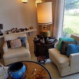 Living room corner with octagonal stand, Oversize lamp, Round coffee table by Baker (finish worn and faded), Swivel chairs, carved fish and duck decoys, and more