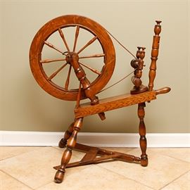 Vintage Oak Spinning Wheel: A vintage oak spinning wheel. This piece features a spoked drive wheel on two turned supports, fixed to one end of a slanted rectangular base. At the other end of the base are the maidens, holding a bobbin. The piece stands on three turned legs, with a treadle and turned support to the bottom.