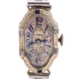 Antique 20K White Gold with Platinum Trim Diamond and Sapphire Wristwatch: An antique 20K white gold wristwatch with platinum trim. This wristwatch features an ornamental hinged case containing four diamonds and two sapphires on an engraved bezel housing a silver tone dial with an engraved center, blue Arabic hour indicators and blue steel hands leading into a peerless open link bracelet. The caseback features a personalized inscription reading “Estelle”.