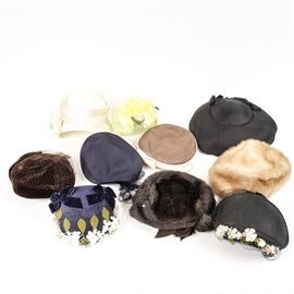 Vintage Hat and Headdress Collection: A vintage hat and headdress collection. Featured in this collection are ten vintage hats in a variety of colors and styles. There are a fur hat marked “McCurdy’s of Rochester Mr. Marc New York”, an unmarked darker colored fur hat, an unmarked black hat with flowers and netting, and an unmarked black hat embellished with black ribbon. Also included are an unmarked navy blue cap with netting, velvet details, and rhinestones, an unmarked cream colored silk hat with a bow, a chocolate brown velvet hat with netting marked “Styled by Bobby Lee, 22”, and a light brown and off-white hat with brown netting and a bow marked “Arlington Hats Dallas”. Finally, there are an unmarked navy blue woven hat with velvet accents, white flowers, and leaves and a light green hat with yellow flowers and netting with a “Union Made GW777459” tag inside.