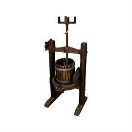 Vintage Wood Wine Press: A vintage wood wine press. The press is composed of a central lidded slatted bucket that sits on a round base with a spout that allows the pressed wine to flow from the bucket to another container after being pressed by the metal press. It is on a stand with two wood square legs joined to their feet by metal brackets.