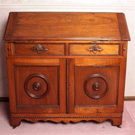 Antique Secretary: An antique solid wood secretary. A rectangular top with slanted front opens to reveal dual shallow drawers with round knobs flanking open shelves in this antique secretary. The desk has two pull out supports to either end with dual drawers above cabinet doors which open to a single shelf. The front of the desk has intricate wooden trim to the drawers and cabinet doors. Lion headed ring pulls of brass are found to the front of both drawers, while the cabinet doors have the same trim with decorative round frames to the center. The desk has a carved apron with bracket feet and rests upon brass castors. Desk is unmarked by maker.