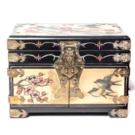 Lacquerware Jewelry Box: A highly ornate lacquerware jewelry box. Comprised of glossy lacquered wood, this box features carved imagery of birds, trees and flowers with gold details and embellished brass accents throughout. the rectangular top opens to a shallow drawer lined in embroidered red silk with a mirror to the reverse of the lid. The front has double doors, with swallows in flight on a golden ground to one side with cherry blossoms on the other, which open to three jewelry drawers in black. Brass plates to the front with basket shaped pulls and brass medallion handles to either side. This box rests upon brass wrapped square feet and is marked to the reverse with a sticker which reads, “Made in Macau.”