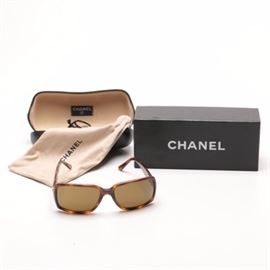 Women's Chanel Tortoise Sunglasses: A pair of women’s Chanel tortoise sunglasses. These sunglasses feature a faux tortoise shell frame with a brown tint to the lenses. The brand logo appears on either side of the temples of the glasses and is also marked to the inside of the right hand arm. These glasses have the serial number BC1931604 and come with a care manual, box, dust cover, and glasses case.
