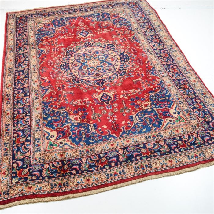 Vintage Hand Knotted Persian Qum Area Rug: A vintage hand knotted Persian Qum area rug. This wool rug features a center medallion in shades of cream, sage, blue, wine and gold on a red, lozenge shaped field. The field is bounded by blue spandrels and surrounded by a blue major border within floral minor borders, and a wine colored guard border marks the perimeter with white cotton fringe at the far ends. This rug is tagged underneath “Made in Iran, Hand Woven Carpet & Rugs…”