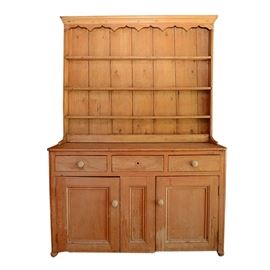 19th Century Pine Stepback Cupboard: An antique pine stepback cupboard. This circa mid to late 19th Century piece features a crown over scalloped crest above open shelving with vertical backing boards, flanked by panel stiles supported by bracket feet. The hutch rests onto a lower dresser with three small drawers including knob pulls and hand cut dovetail joinery over a pair of recessed panel front doors with knob pulls. The doors open to reveal slatted shelving and the unit terminates in extended panel feet.