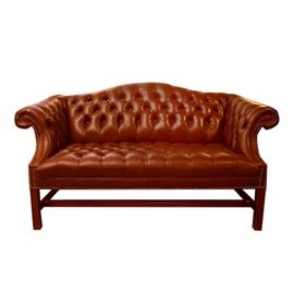 Contemporary Leather Settee: A contemporary leather settee. This camelback settee with button tufted back and seat is flanked by scrolling armrests with nailhead trim detail to the supports and apron front. The chair sits on a wooden frame with straight legs joined by an H-stretcher. There are no visible maker’s marks.
