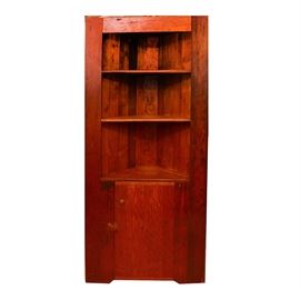 Pine Corner Cabinet: A pine corner cabinet. This vintage cabinet features a triangular footprint with three open shelves above a panel front cabinet door including swing latch and knob pull. The cabinet includes panel stiles, a vertical slat back and is finished in a cherry stain.