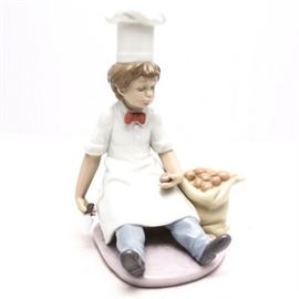 Lladró "Chef's Apprentice" Figurine #6233: A Lladró figurine titled Chef’s Apprentice, #6233. The young chef is sitting on the floor peeling potatoes with a potato in his right hand and a sack of potatoes to his left. The young chef is dressed in an apron, chef’s hat and red bow tie. The piece was sculpted by Antonio Ramos, released in 1994 and retired in 1998. The piece is marked to the underside, “Lladró hand made in Spain Daisa 1994” and the bellflower.