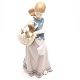 Lladró "Little Dogs on Hip" Figurine #1311: A Lladró figurine titled Little Dogs on Hip, #1311. The figurine depicts a young girl with a long lavender skirt and blue top. She is holding a basket of puppies on her hip; one puppy is looking up at her as the other two are in the basket. The figurine was issued in 1974 and retired in 1992; originally sculpted by Juan Huerta.