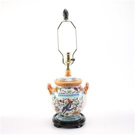 Orange Accented Blue Floral Ceramic Table Lamp: An orange accented blue floral ceramic table lamp. The lamp has a black flame finial and gold tone harp with a lid style vase cap with orange trim and a ceramic body with a white finish that showcases a richly colored blue, orange red and green floral, fruit and bird design. The light also features orange rope style side handles and a round bevel edged wood base with a black lacquered finish and bracket feet.