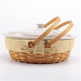 Longaberger Basket With White Ceramic Oval Casserole Dish: A Longaberger basket with a white ceramic casserole dish. This basket is woven from wooden strips and has two looped handles, with a tan cloth liner. The basket comes with an oval-shaped casserole dish with a lid which fits snuggly inside. The underside of the basket is marked “Longaberger Baskets Handwoven Dresden, Ohio”.