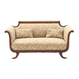 Regency Style Sofa: A Regency style sofa. This sofa has reeded rails, scrolled arms, and curved, reeded legs with brass tone paw feet. It is covered in a light floral upholstery and comes with two matching pillows.