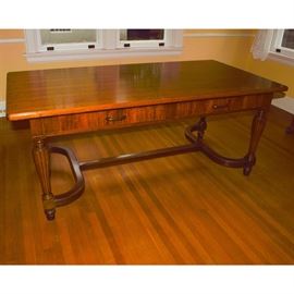 Library Trestle Table: A library trestle table. Constructed of mixed hardwoods and veneers, the table features a top with ogee fillet edges, radius corners, and a generous overhang. The apron houses two aligned drawers with dovetail joinery and patinated pulls. It is raised by reeded and tapered legs with an arched trestle-style base. The top is detached for easier transport. This item is located on the main level of the home and will require assistance in moving.