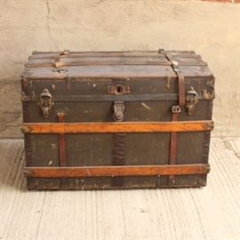 Antique Steamer Trunk: An antique steamer trunk. This unit features a pull our sectioned shelf for securing smaller items, with a large bottom section for folded clothing. The exterior features Art Deco style wood banding, a key lock, two flip down closures, and leather securing belts. No key is present and trunk does not lock.