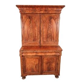 Antique Empire Style Flame Mahogany Secretary: An antique Empire style mahogany secretary. This nineteenth-century secretary is made of flame mahogany and other woods, featuring substantial crown molding at the top, above a cabinet with matched mahogany veneer on the two Gothic, recessed paneled doors with beaded trim. The cabinet is fitted with brass key holes, and opens to reveal a variety of divided compartments, adjustable shelves and two small drawers fitted with round brass knobs. The hinged desktop folds down and rests on pull-out supports with rounded corners that match the corners of the secretary. There is an additional cabinet at the bottom, with recessed paneled doors, which also have beaded trim, and the secretary rests on bun feet. No maker’s marks. This heavy item is located in a basement that has a door leading to the outside, however buyer should be aware that the path from this door to the street is up a hill.