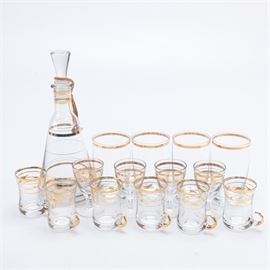 Vintage Glass Decanter With Glasses: A vintage glass decanter with glasses. This set of fifteen pieces includes a decanter, six small handled glasses, four small tumblers, and four stemmed glasses. These pieces feature a clear glass body with gold tone detail throughout. There are no visible maker’s marks.